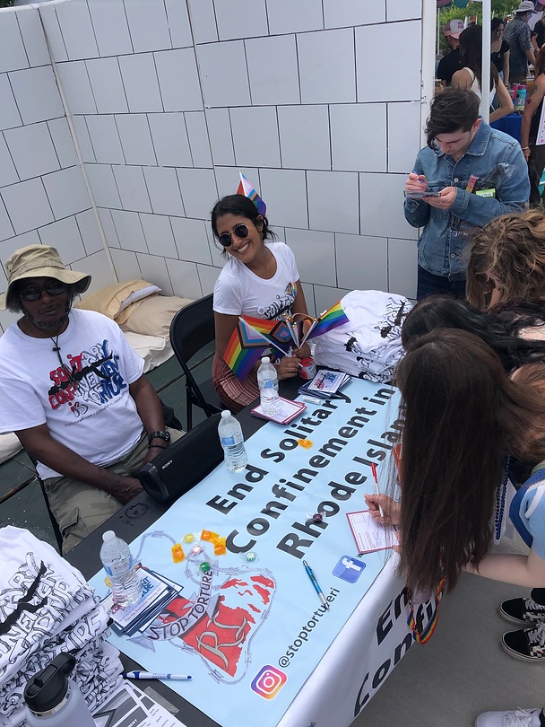 Two people sit behind a table that says, "End Solitary Confinement in Rhode Island." They are wearing "Solitary confinement is torture t-shirts. They sit in front of a mock solitary confinement cell. People gather around signing post cards.