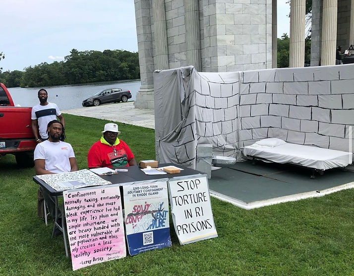 Three people sit behind a table with signs that say, "Stop Torture in RI prisons" and other messages. A mock solitary confinement cell is next to them.