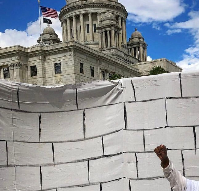 A person sits in front of the RI State Housein our life-size mock solitary confinement cell. His fist is in the air.